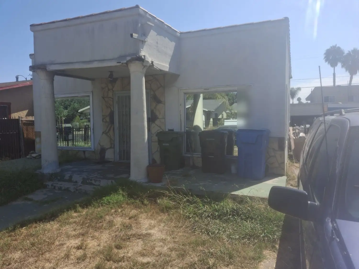 The house: a stucco front with a columned porch, bins lined up and the yard overgrown