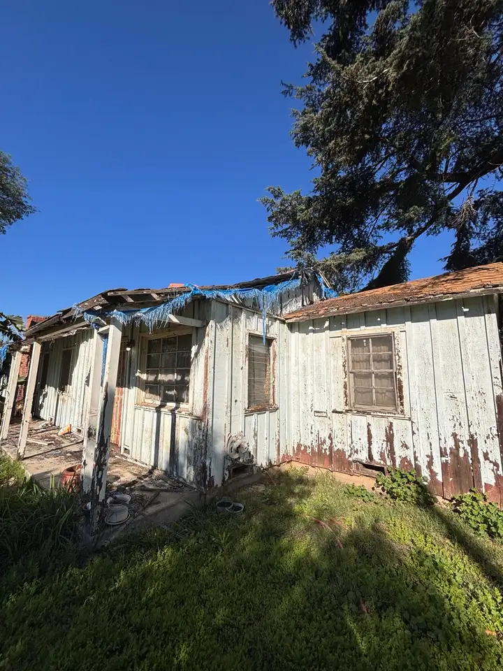 The side of the house, paint peeling and the roof edge torn open